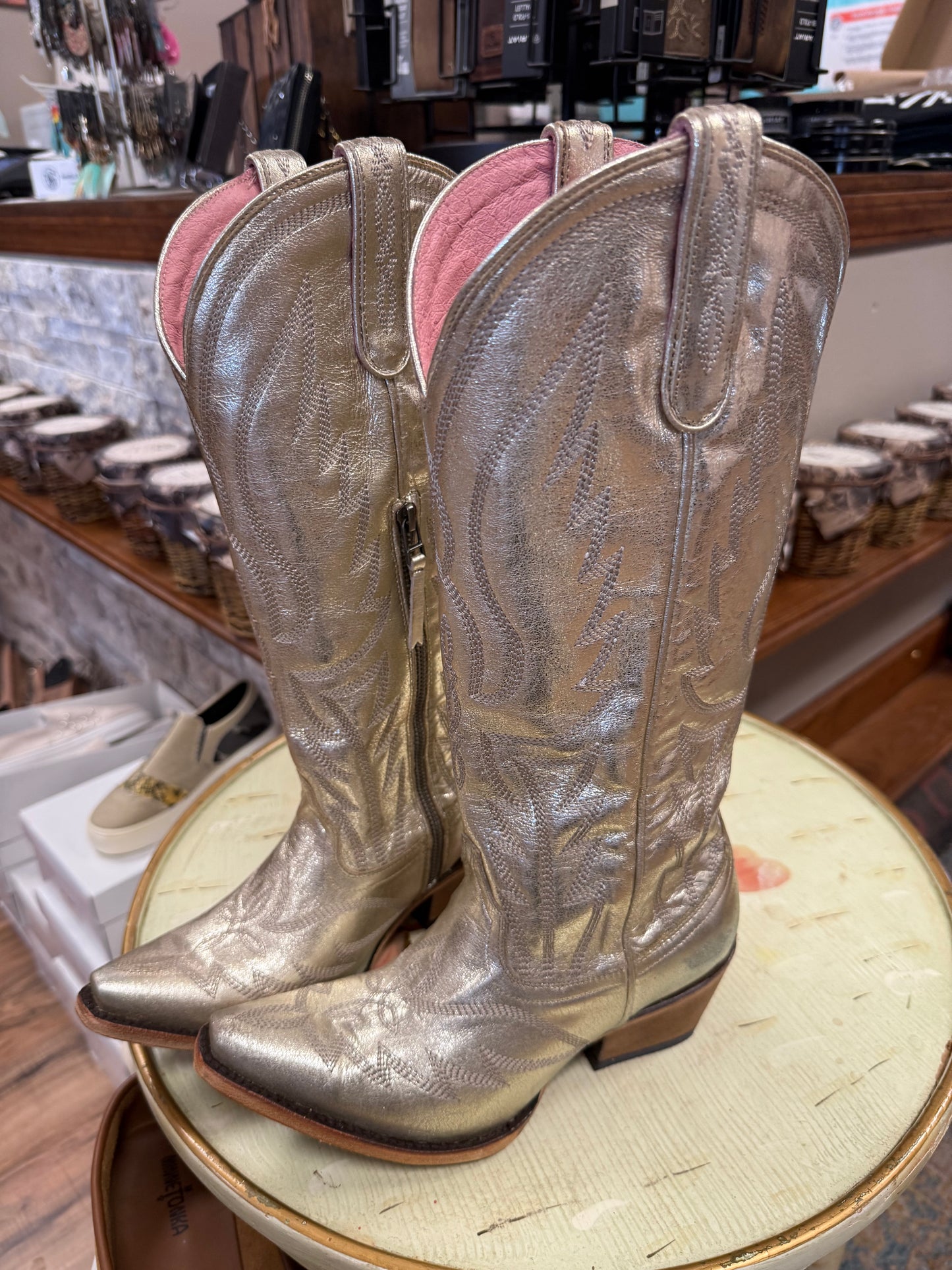 Pair of metallic silver cowboy boots on a round surface with a store interior in the background.