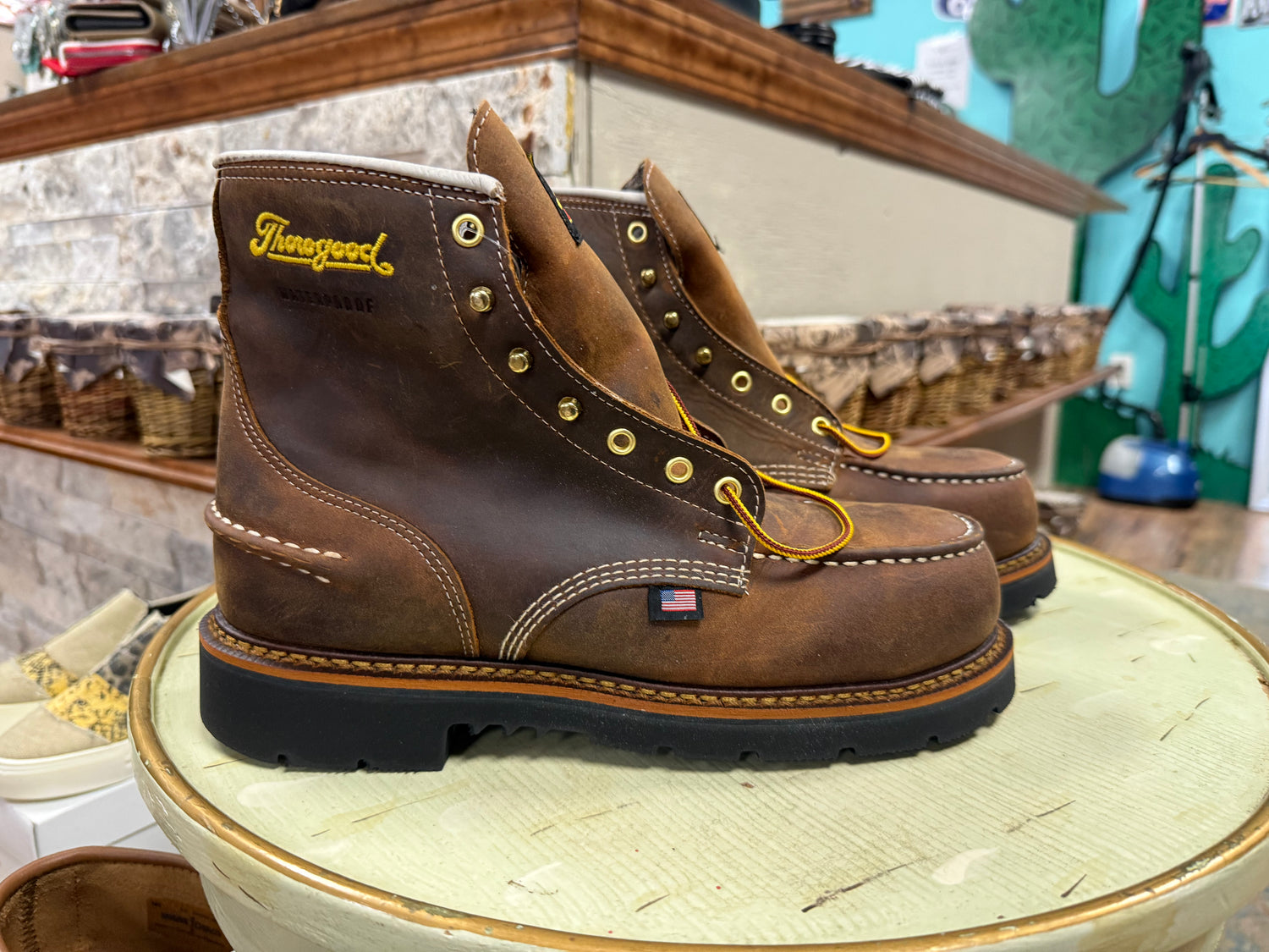 Pair of brown work boots with gold accents on a wooden stool in a store setting.