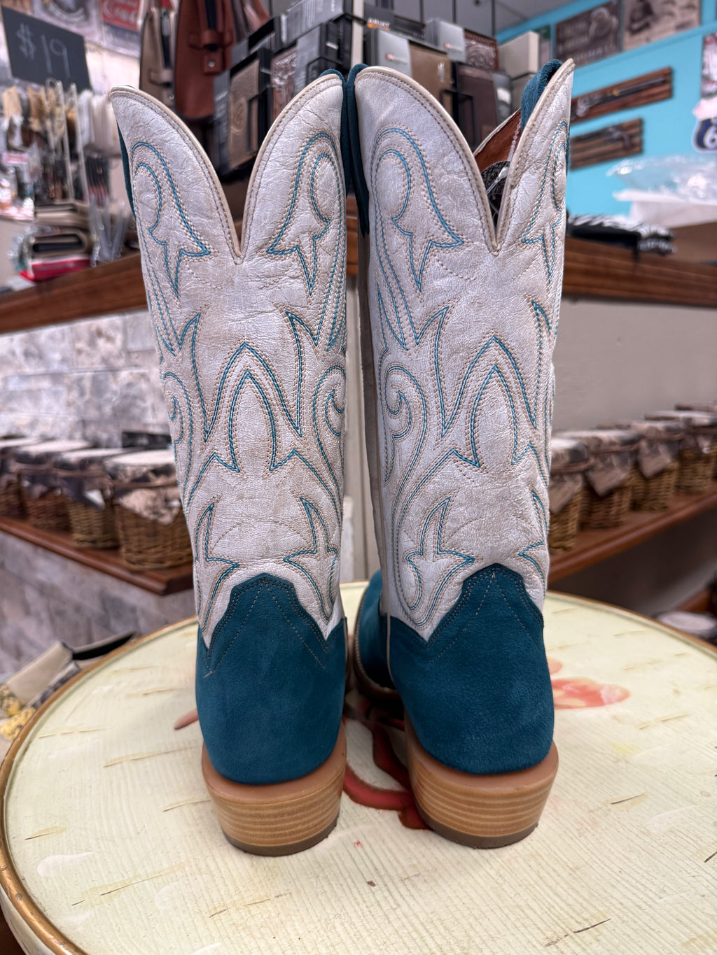 Pair of white cowboy boots with blue soles on a round surface in a store setting.