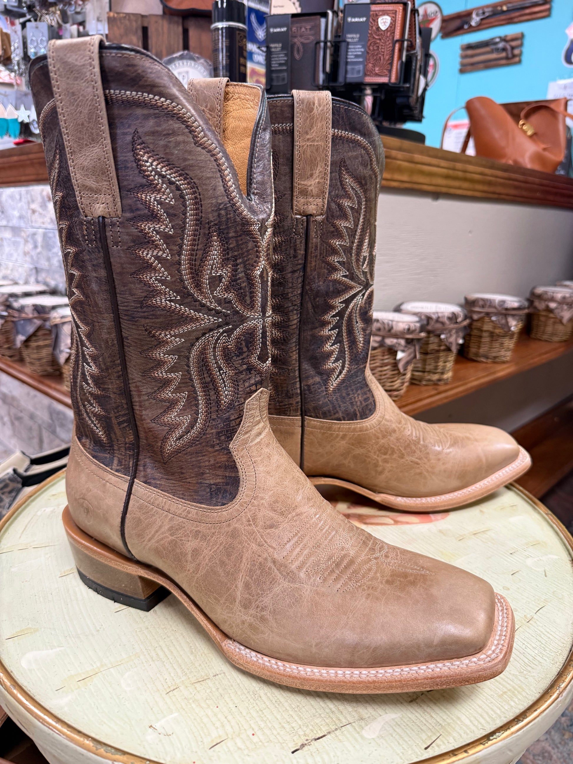 Pair of brown and beige cowboy boots on a white surface with a store interior in the background.