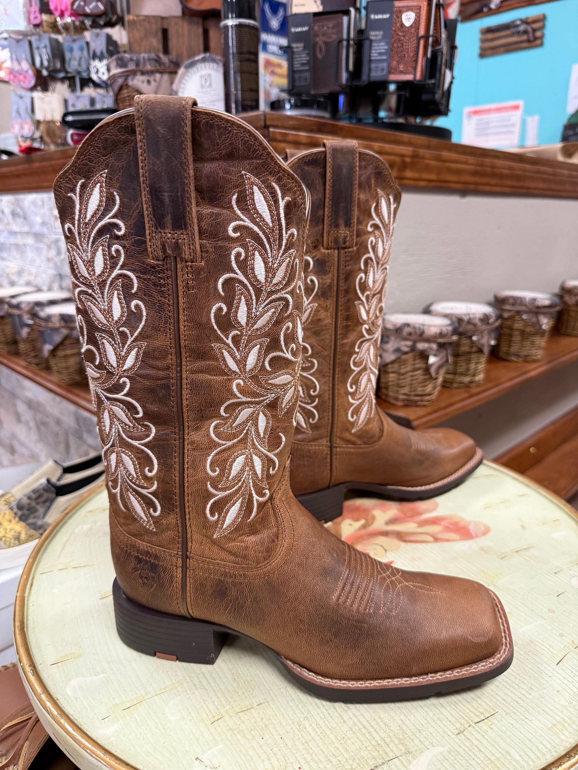 Brown cowboy boots with white embroidery on a round surface in a store setting.