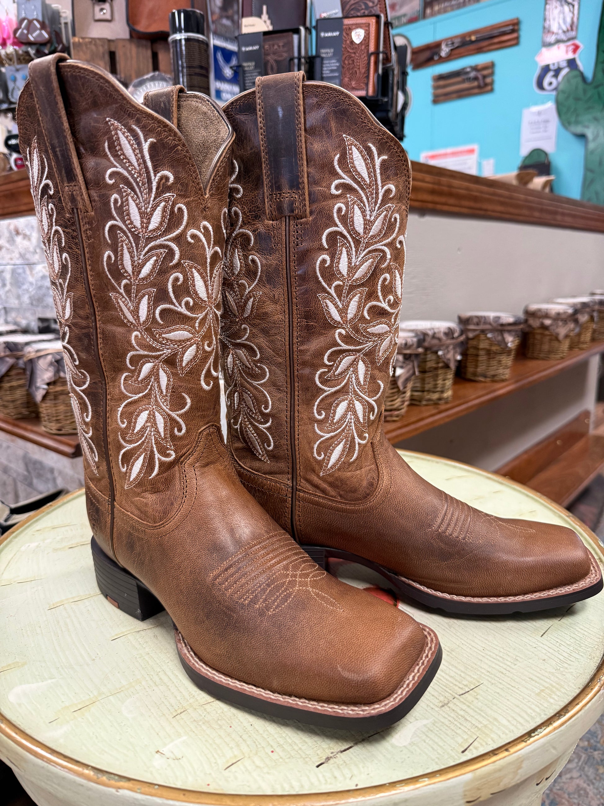 Brown cowboy boots with white embroidery on a round surface, with a store setting in the background.