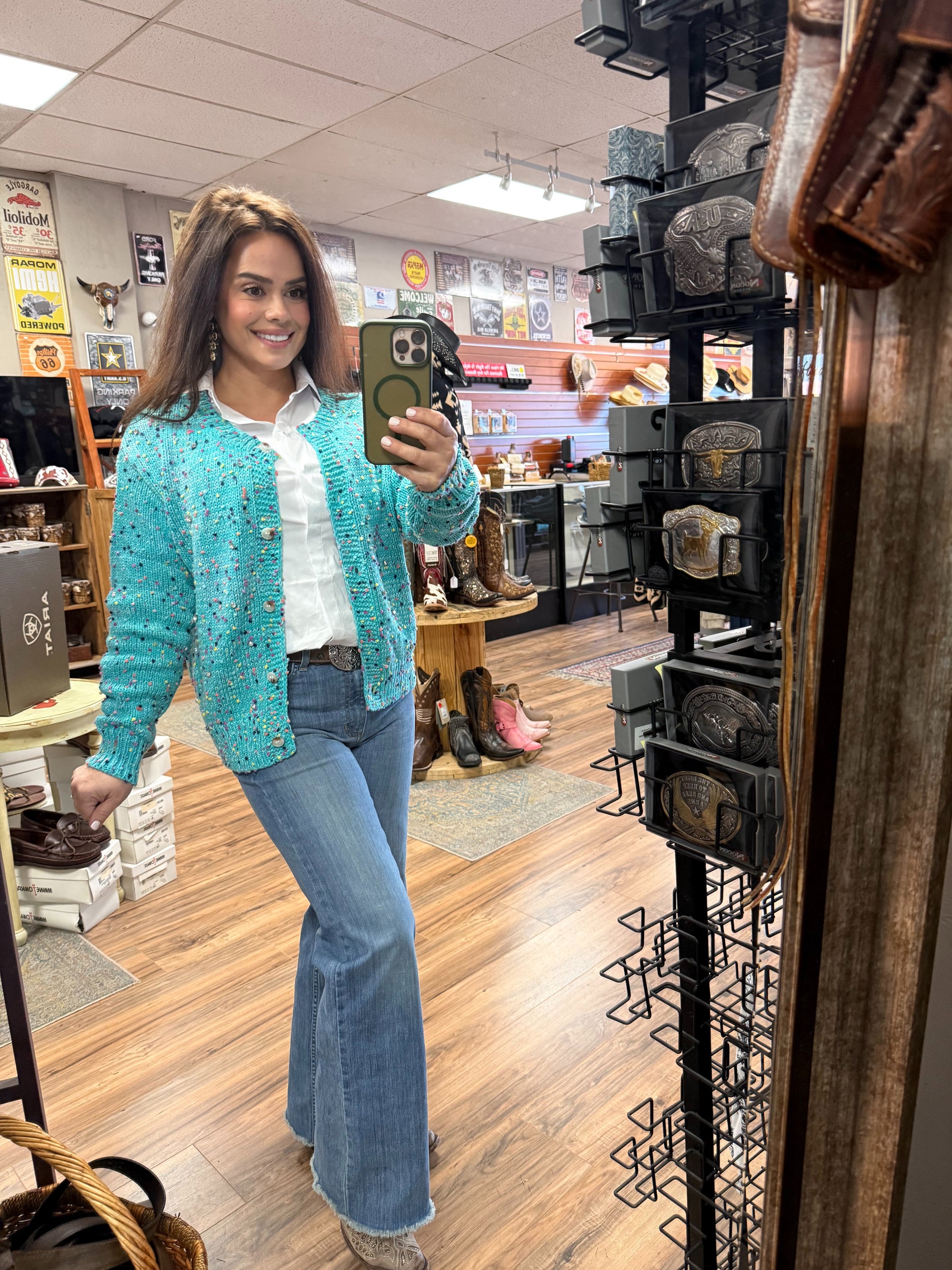 Woman taking a mirror selfie wearing a teal cardigan and jeans in a store.