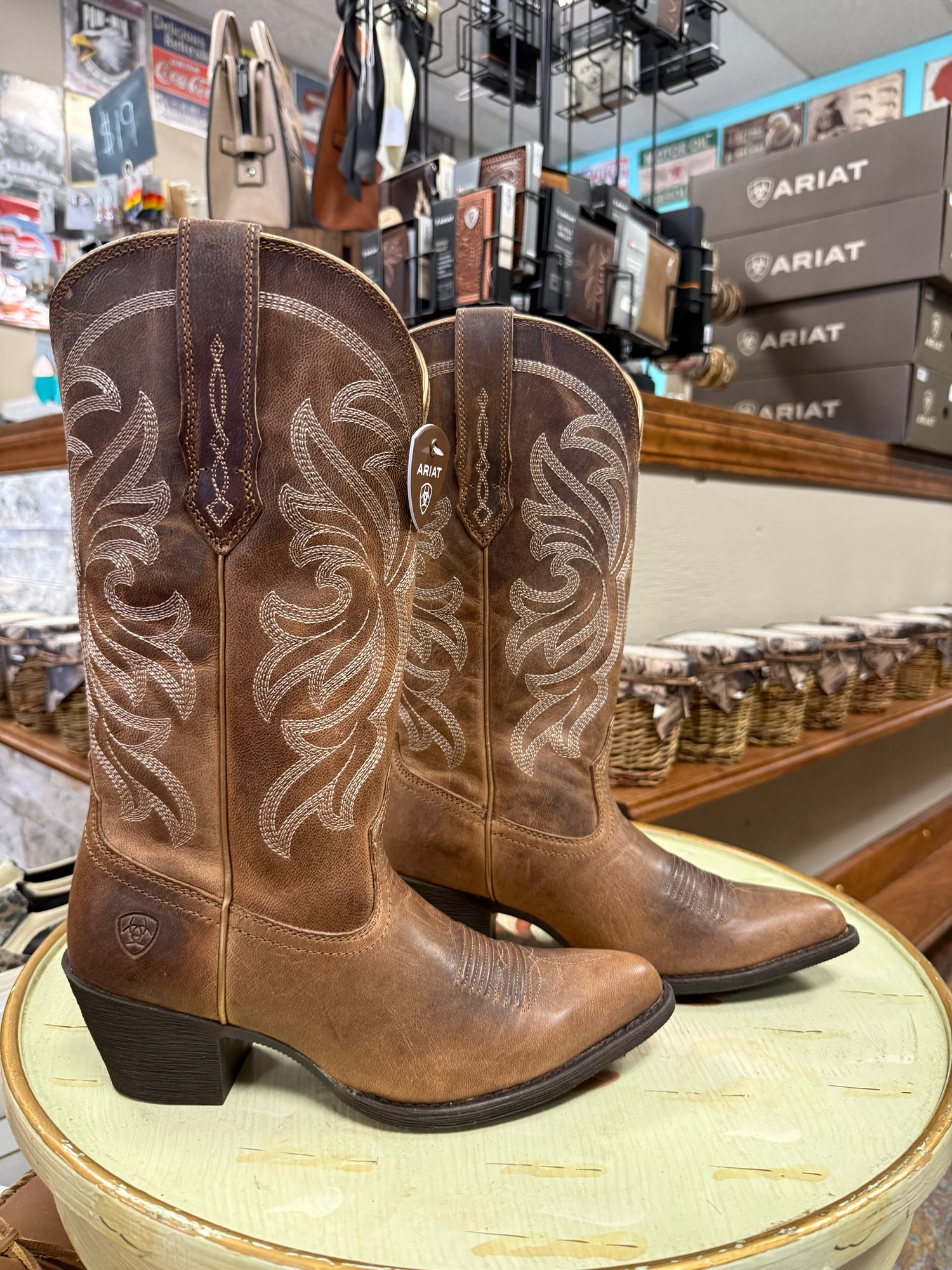 Brown cowboy boots with intricate white stitching on a round surface in a store setting.