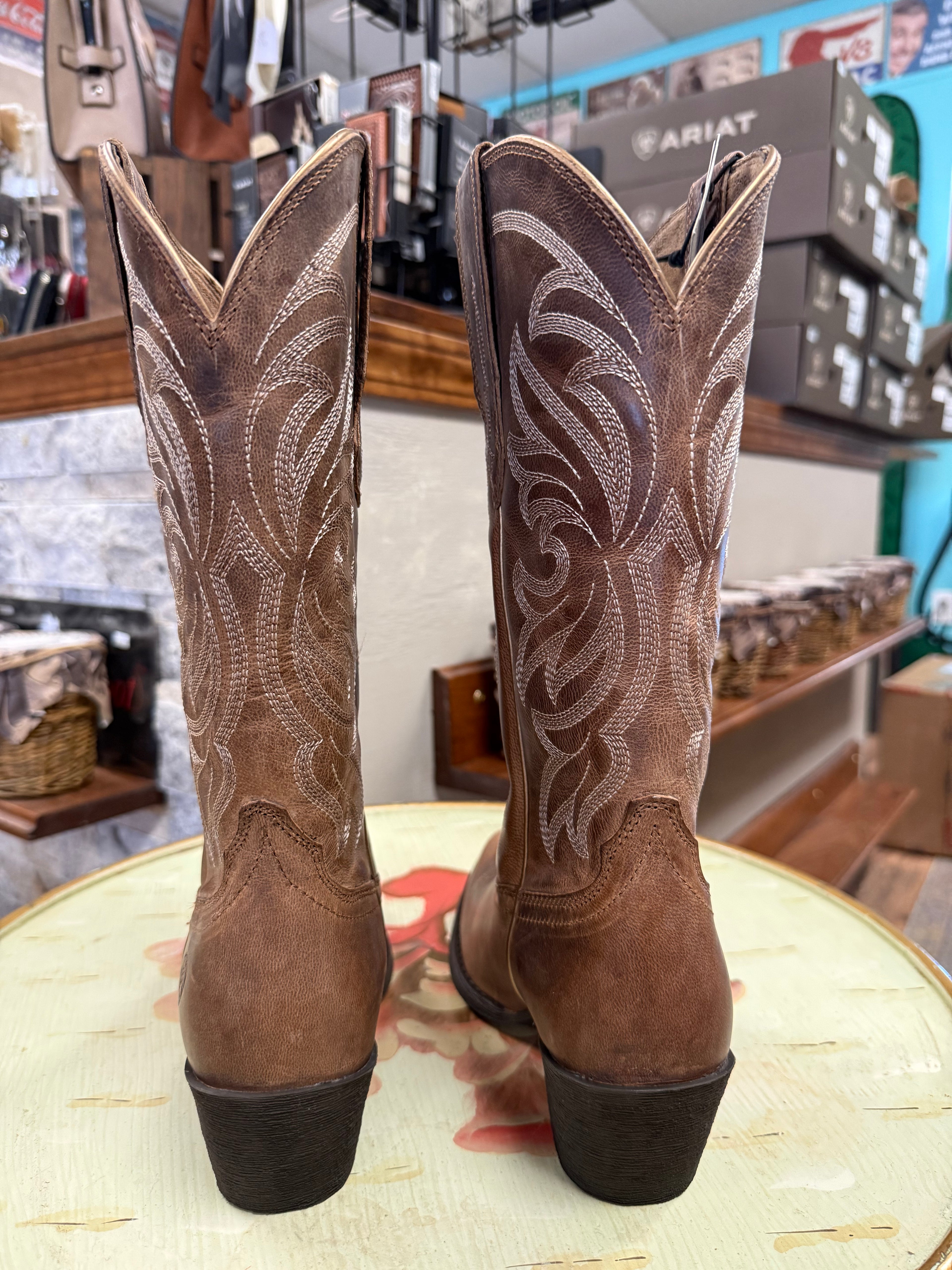 Pair of brown cowboy boots with intricate white stitching on a round surface, store interior in the background.