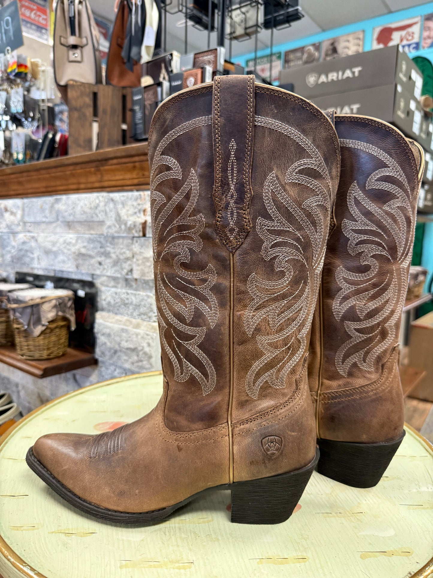 Pair of brown cowboy boots with intricate designs on a round surface in a store setting.
