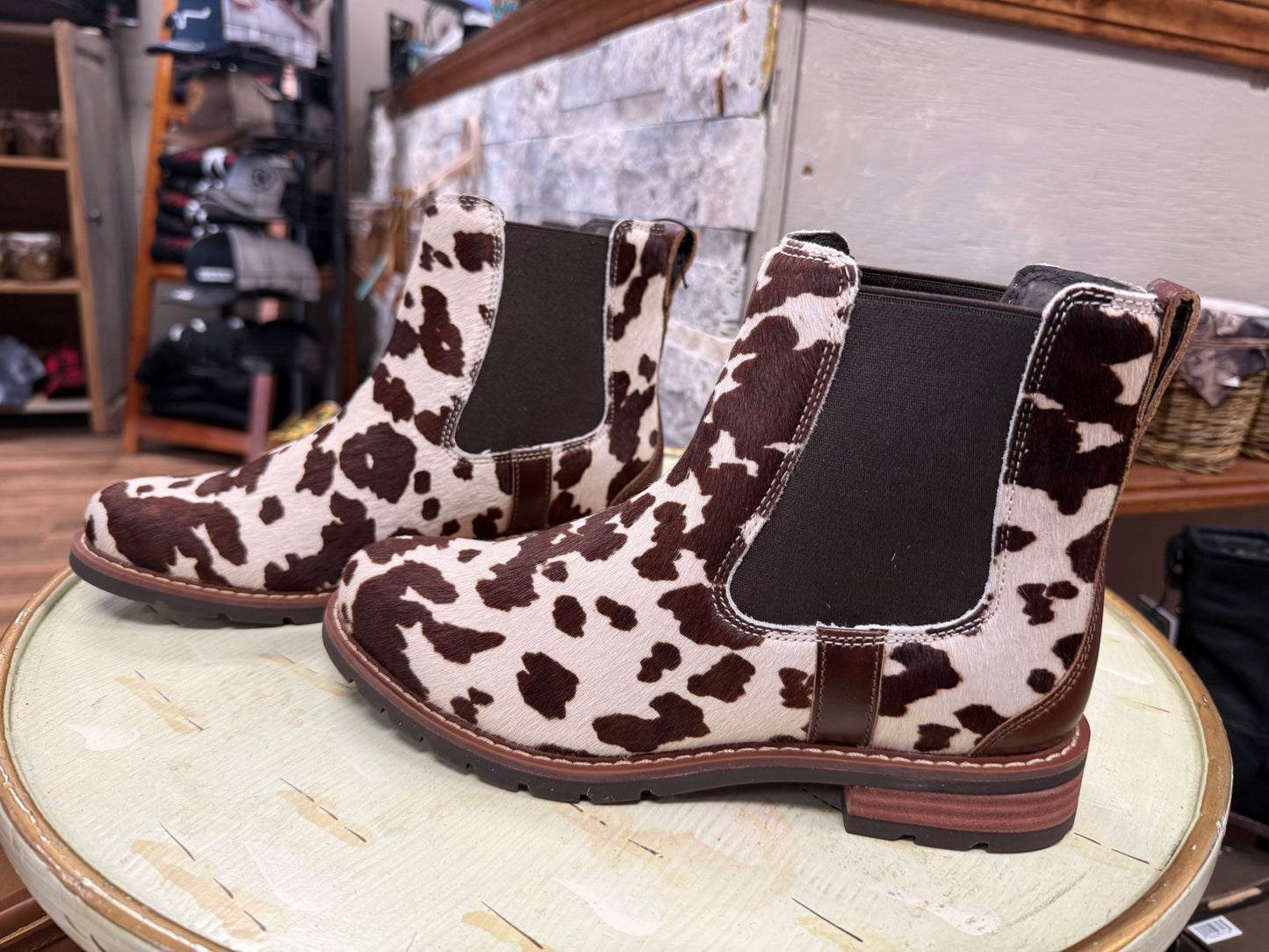 Cow print Chelsea boots on a round wooden table with a store interior background.