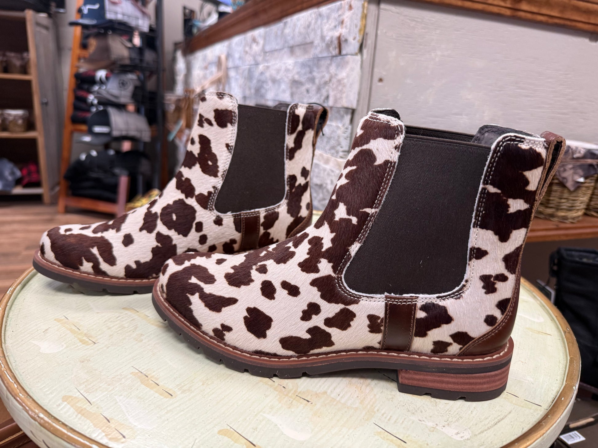 Cow print Chelsea boots on a round wooden table with a store interior background.
