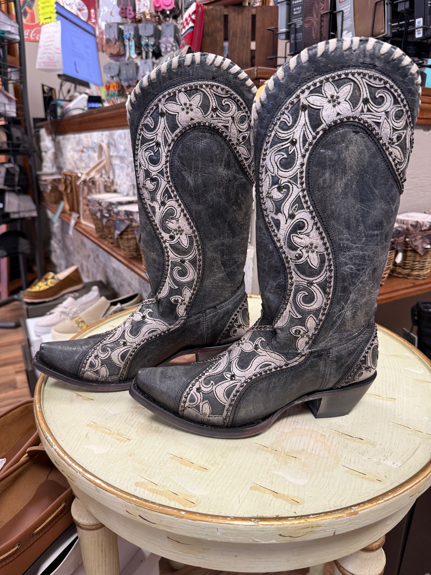 Pair of gray cowboy boots with white embroidery on a round table in a store setting.