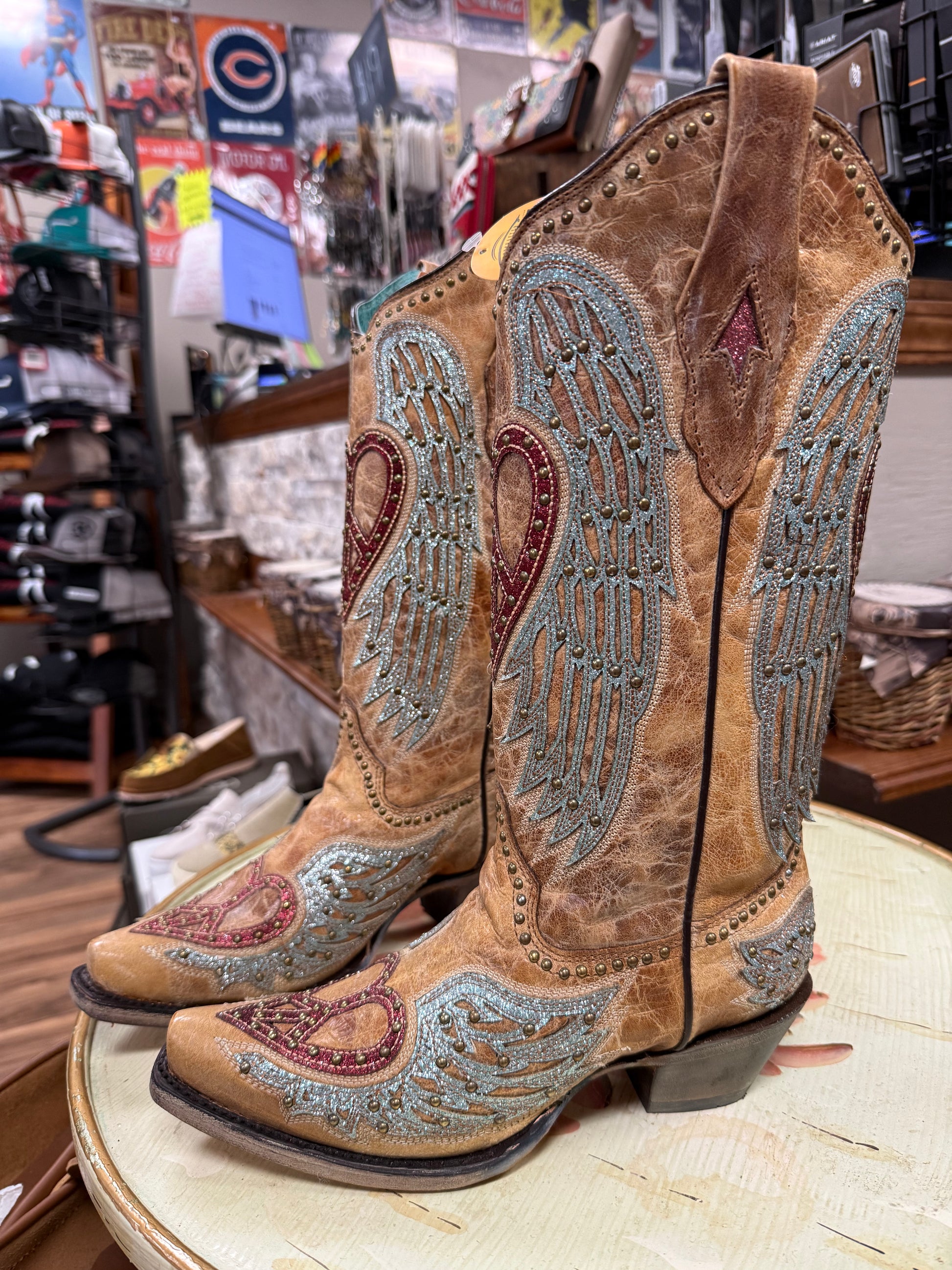 Pair of brown cowboy boots with decorative wings and hearts on a round table in a store setting.