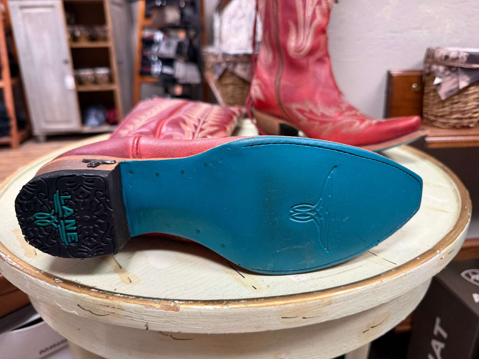 Close-up of a blue sole with a brand logo on a wooden stool, with red cowboy boots in the background.