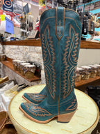 Pair of teal cowboy boots with brown leather stitching on a round table in a store setting.