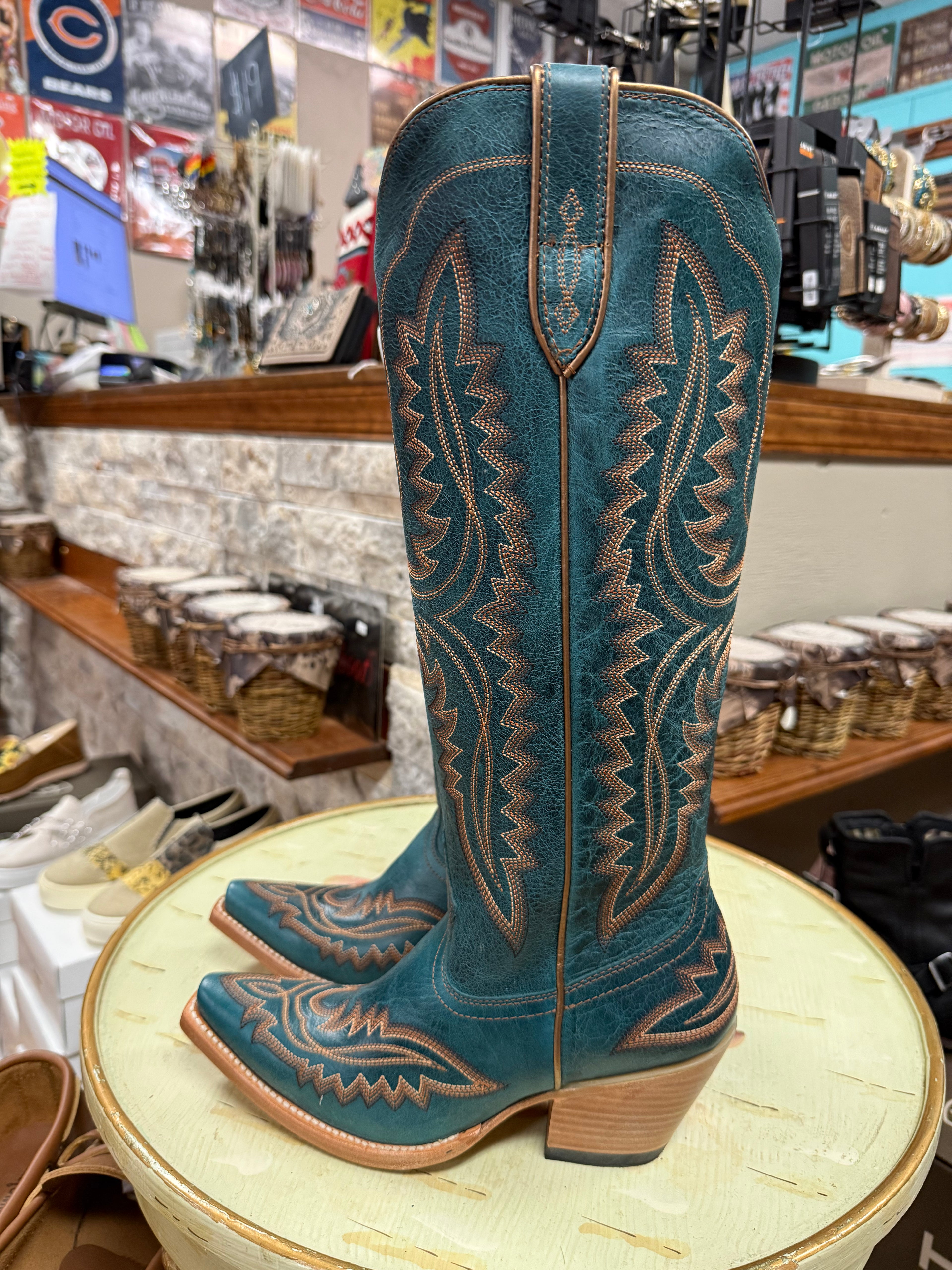 Pair of teal cowboy boots with brown leather stitching on a round table in a store setting.