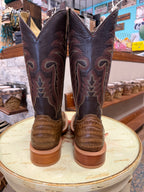 Pair of brown cowboy boots with intricate patterns on a round wooden stool in a store setting.