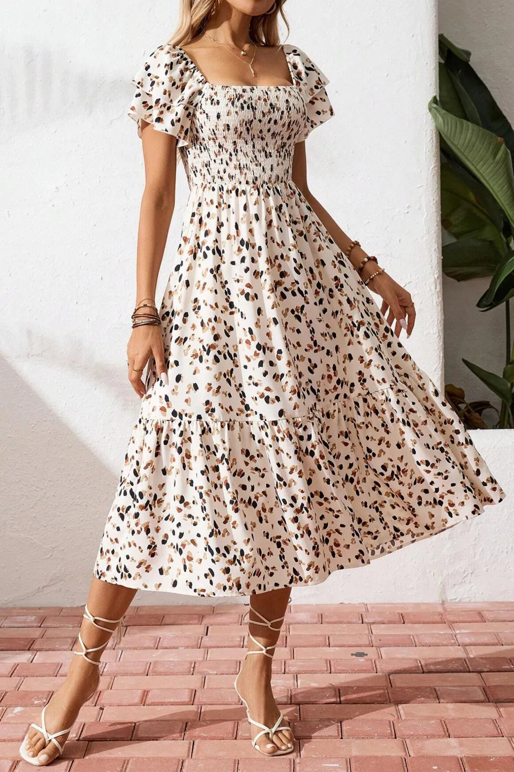 Woman wearing a floral dress standing against a white wall with a plant in the background.
